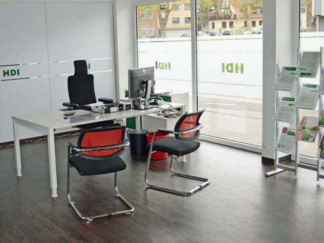 In dem Büro steht ein Schreibtisch auf dem ein PC steht. Hinter dem Schreibtisch steht ein schwarzer Stuhl und vor dem Tisch stehen zwei rote Stühle. Rechts in der Ecke steht ein Aufsteller mit HDI Zeitschriften. Außerdem sieht man eine Fensterfront