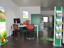 Rechts und Links sind HDI Aufsteller, der rechte ist mit Zeitschriften befüllt. Dahinter steht ein Tisch mit PC, vor und hinter dem Tisch stehen Stühle. Hinter dem Tisch steht ein grünes Regal und ein Poster hängt an der Wand. Links vom Regal steht ein weiterer Aufsteller und daneben geht ein Gang weiter nach hinten