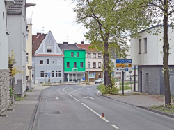 grünes Haus mit HDI Logo, Fenster und unten ist eine Fensterfront, links neben der Agentur steht ein graues Haus und rechts neben der Agentur steht ein rosa, braunes Haus. Vor den Häusern ist eine normale graue Straße mit Fahrmarkierungen