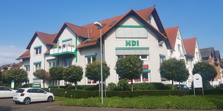 Die Agentur befindet sic in einem weißen Gebäude mit rotem Dach. Man erkennt über einem Fenster das HDI Logo. Vor dem Gebäude stehen grüne Bäume und grüne Büsche. Außerdem scheint ein blauer Himmel.
