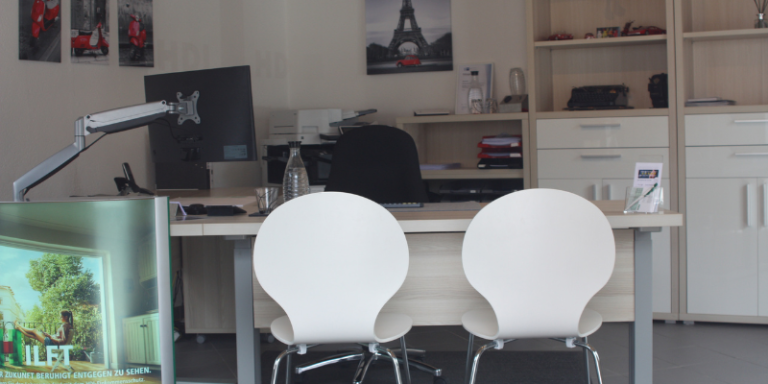 HDI Büro: Man sieht einen Schreibtisch und dahinter eine Regalwand, über einem der Regale hängt ein Bild vom Eifelturm, an der linken Wand hängen drei weitere Bilder mit roten Akzenten, vor dem Schreibtisch stehen zwei Stühle die für Beratungen gedacht sind, vorne links im Bild sieht man eine Werbeaufsteller von HDI