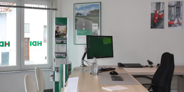 HDI Büro: man sieht einen Schreibtisch auf dem sich u.a. eine PC und PC-Zubehör befindet, Büroartiekl und eine Wasserflasche, auf der einen Seite des Tisches steht ein schwarzer Schreibtischstuhl, auf der anderen Seite stehen zwei weiße Stühle, an der Wand im Hintergrund hängen mehrere Bilder und man sieht mehrere dekorative Werbeelemnte von HDI