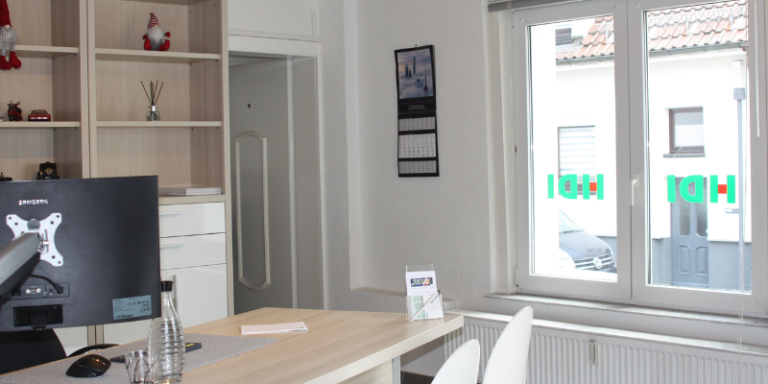 HDI Büro: Man sieht einen Schreibtisch und dahinter eine Regalwand,  vor dem Schreibtisch stehen zwei Stühle die für Beratungen gedacht sind, neben der Regalfront ist eine geöffnete Tür, die in ein angrenzendes Zimmer führt, man sieht Fenster mit dem HDI Logo und links neben den Fenster hängt ein Fotokalender an der Wand