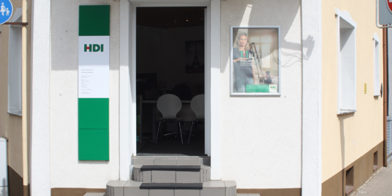 Man sieht den Eingang zum HDI Büro von der Straße aus, die Tür steht offen und gibt den Blick frei auf einen Schreibtisch vor dem zwei Stühle stehen, rerchts neben der Tür hängt eine Werbeplakat von HDI, links neben der Tür hängt ein Schild mit dem HDI Logo und Informationen über die Agentur