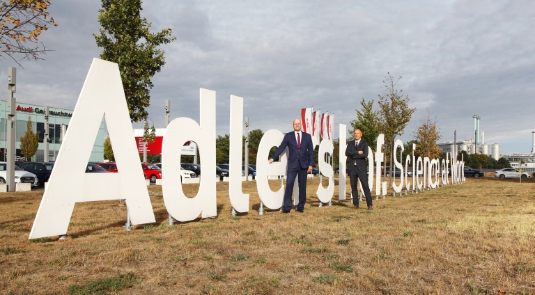 Michael und Gleitz und Mitarbeiter vor großem Schriftzug in Form von haptischen, mannsgroßen Buchstaben auf einer braunen Wiese, die Buchstaben stellen das Logo von Adlershof - Science at Work da