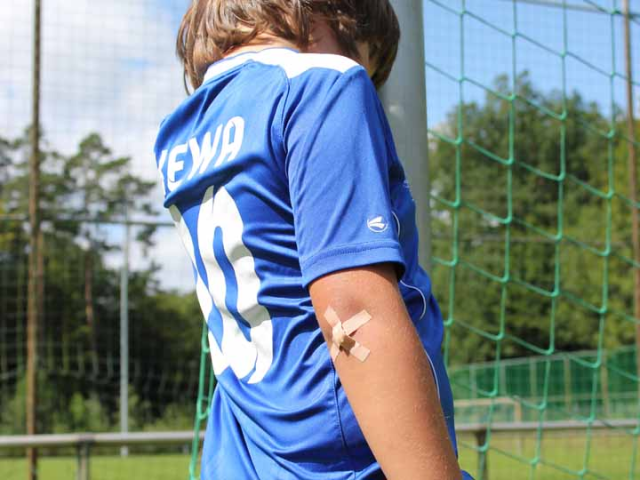 ein Kind mit braunen Haare wurde von der Seite fotografiert. Das Krind hat ein blaues trikot an und auf seinem Rücken steht in weiß KEWA und eine Zahl. An seinem Ellenbogen hat das Kind ein Pflasterkreuz. Das Kind steht vor einem Tor, hinter dem Tor ist ein grüner Platz zusehen und dahinter ein Wald mit grünen Bäumen. Außerdem ist ein blauer himmel zusehen.