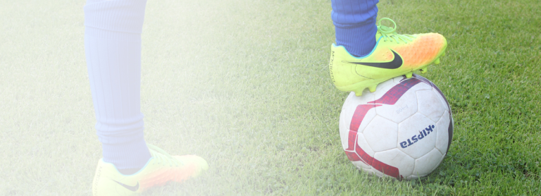 Man sieht ein weiß roten Ball. Auf dem ein gelb orangerner Fußballschuhe steht. Der Ball liegt auf einem grünen Rasen.