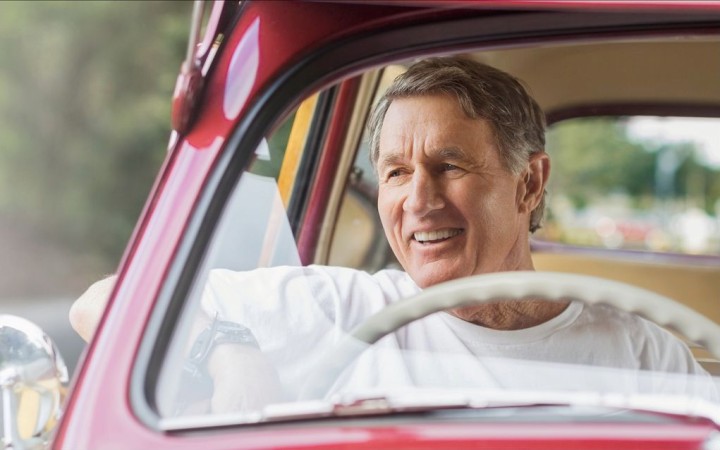 Älterer Mann in einem roten Oldtimer mit dem Lenkrad auf der rechten Seite, hat den Arm lässig aus dem Fenster gelehnt und lächelt in den Außenspiegel