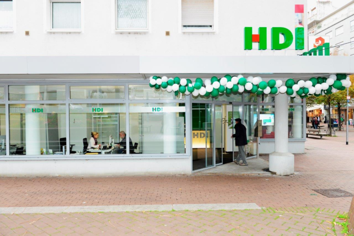 Man guckt von der Straße auf ein Hochhaus, im Erdgeschoss befindet sich die HDI Agentur, das Vordach ist mit grünen und weißen Luftballons geschmückt, ein Mann geht gerade durch die Eingangstür, durch viele bodentiefe Fenster erhält mein einen Blick auf die Innenräume der Agentur, wo gerade u.a. ein Beratungsgespräch zwischen einem Mann und einer Frau statffindet