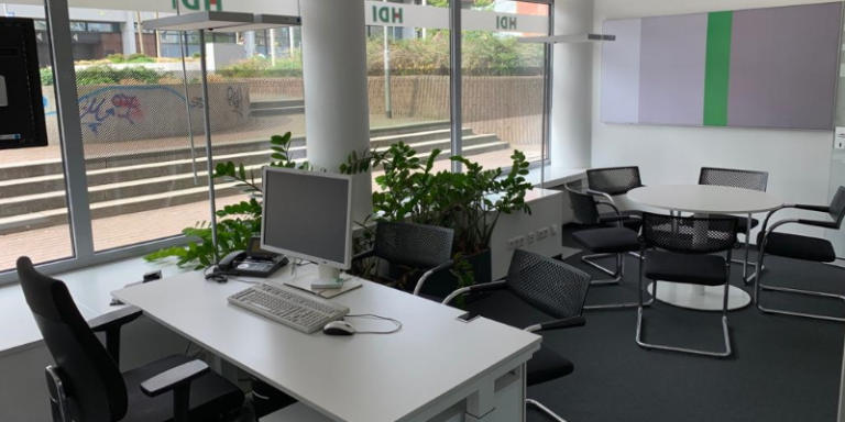 Man sieht ein Büro, im Vordergrund befindet sich ein Schreibtisch mit Stuhl, auf dem Schreibtisch steht ein PC und PC-Zubehör sowie ein Telefon, vor der Fensterfront befindet sich eine Bank und Säulen, von denen eine von Zimmerpflanzen umstellt ist, im hinteren Teil des Raums befindet sich ein runder Tisch mit fünf Stühlen