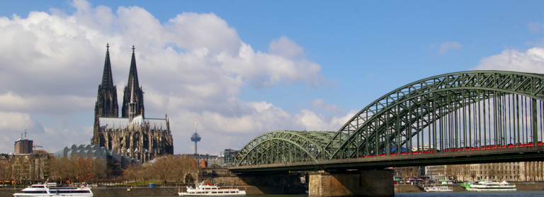 Blick auf Köln, zu sehen sind die Hohenzollernbrücke, der Kökner Dom und der Rhein auf dem Schiffe unterwegs sind. Der Himmel ist zum Teil bewölkt.