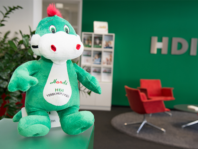 grün, weißer HDI Kuscheltier Drache. Im Hintergrund steht links von dem Kuscheltier eine grüne Pflanze und rechts von dem kuscheltier steht ein weißes Regal mit Zeitschriften.