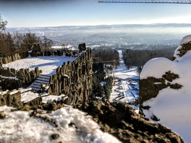Natur im Winter fotografiert mit Schnee. Man sieht eine Aussichtsplattform und eine Treppe. wenn man runter guckt, sieht man Berge, Schne, einen blauen Himmel und kahle braune bäume.