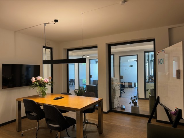 In der Mitte steht ein brauner Tisch, um den Tisch stehen vier schwarze Stühle und auf dem Tisch steht ein Blumestrauß. Links vom Tisch ist ein Fernseher an der Wand und hinter dem Tisch ist eine Fensterfront, durch die man in den Flur gucken kann. Rechts neben dem Tisch steht ein Whiteboard. 