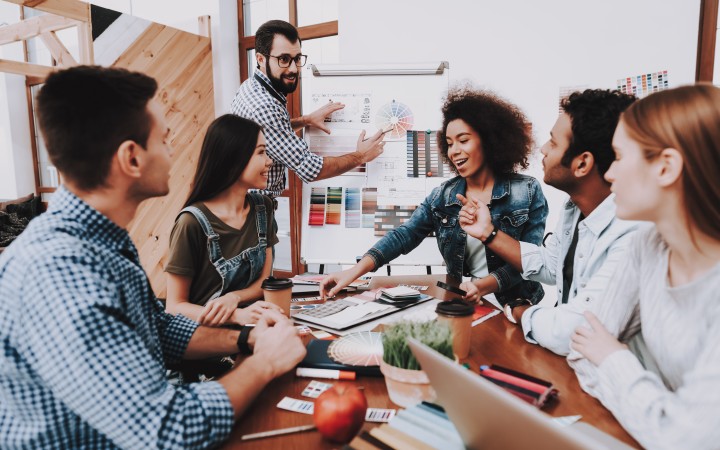 Das Bild zeigt ein sechs-köpfiges Team an einem Tisch sitzend, wobei einer aus dem Team vorne an einem Flipchart steht und etwas erklärt. 
