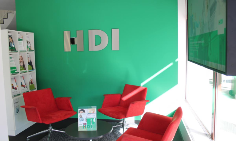 In dem Büro stehe drei rote Stühle, diese stehen um einen runden Glastisch. Auf dem Glastisch steht ein kleiner Aufsteller mit HDI Flyern. Diese Möbel stehen vor einer grünen Wand, an dem das silberne HDI Logo hängt.