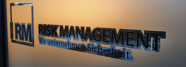 Schriftzug "RISK MANAGEMENT - Wir unternehmen Sicherheit" auf einem Milchglas-Fenster.