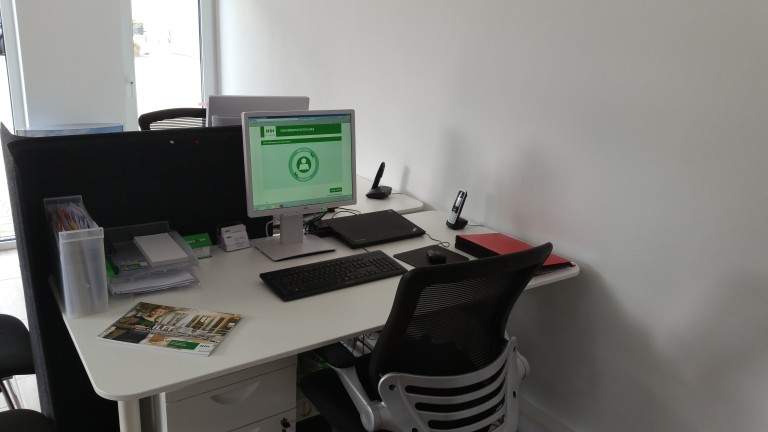 Auf einem weißen Schreibtisch steht ein Monitor, auf dem ein HDI internes Programm geöffnet ist. Daneben befindet sich ein Laptop. Außerdem befinden sich eine Tastatur und Werbepospekte auf dem Tisch.