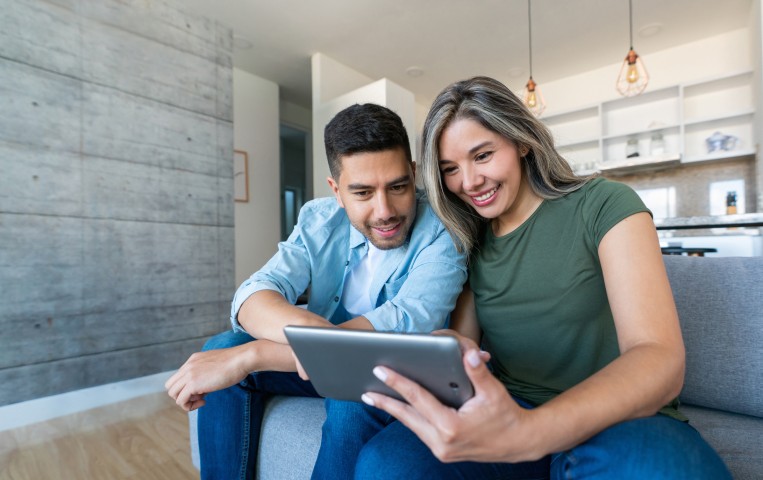 Ein Paar mit Tablet sitzt auf der Couch und schaut gemeinsam auf ein Tablet. Sie lächeln beide, das Bild soll eine Online-Beratung nachstellen.
