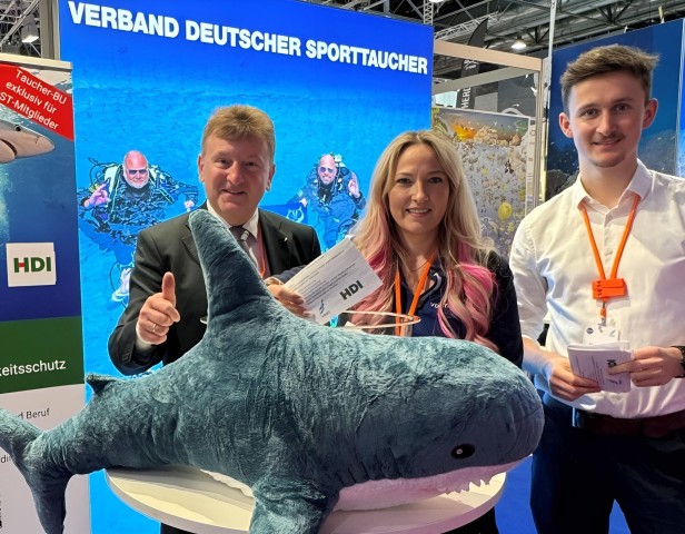 Dörfler und zwei Mitarbeiter befinden sich auf der Messe an dem Stand Verband Deutscher Sporttaucher und vor ihnen liegt ein Hai-Kuscheltier