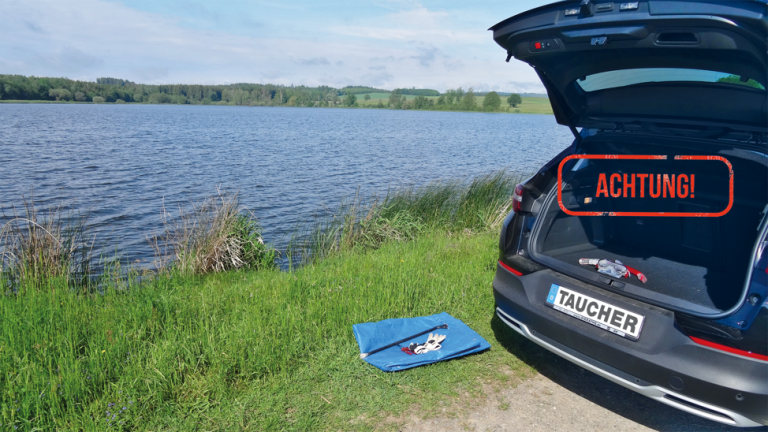 Man sieht das Heck eines Autos. Der Kofferraum steht offen und darüber steht in roten Großbuchstaben das Wort "ACHTUNG!". Auf dem Kennzeichen des Autos steht Taucher. Im Kofferraum liegt ein Schnrochelset. Neben dem Kofferraum liegt auf dem Gras eine blaue Plane. Das Auto parkt einem gras- und schilfbewachsenen Seeufer.