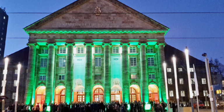 Man sieht ein Gebäude mit großen Säulen und vielen Menschen davor. Zwischen den Säulen hängen Banner mit dem HDI Logo. Das Gebäude wird grün beleuchtet.