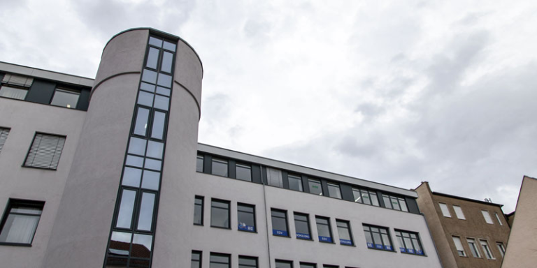 Man sieht ein graues Gebäude mit vielen Fenstern. Ein Teil des Gebäudes ist rundlich.