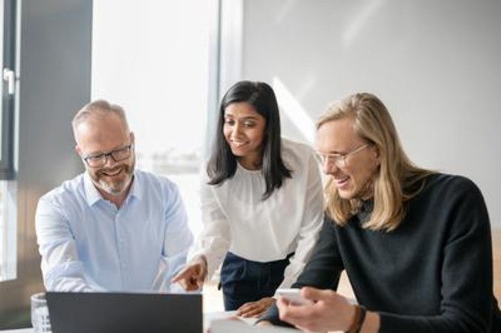 Drei Mitarbeiter sitzen an einem Tisch auf dem ein Laptop steht. Links sitzt ein Mitarbeiter, der lächelnd auf den Laptop guckt. Rechts neben dem Mitarbeiter ist eine Mitarbeiterin, sie steht am Tisch und zeigt lächelnd mit dem Finger auf den Laptop. Rechts neben der Mitarbeiterin sitzt ein Mitarbeiter, der lächelnd auf den Laptop guckt. 
