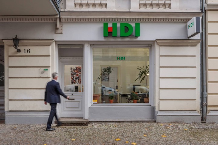 die Agentur befindet sich in einem beigen Haus. Man erkennt ein fenster, durch dieses sieht man ins Büro. Über diesem Fenster befindet sich das HDI Logo und links neben dem Fenster ist eine Glatür, woe gerade ein Mann drauf zu läuft. Vor der Agentur ist ein grauer Gehweg