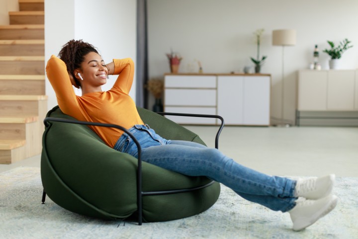 Eine Frau sitzt entspannt in einem grünen Sitzsack auf einem weißen Teppich. Hinter Ihr erkennt man ein weißes Regal, eine Lampe und ein weiters weißes Regal