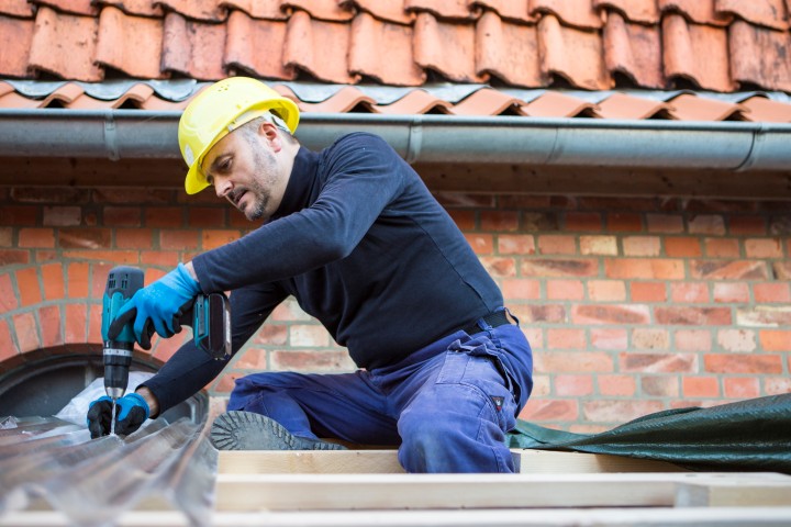 Der Mann sitzt auf einem Dach und bohrt mit einer Bohrmaschine Nägel in das Dach. 