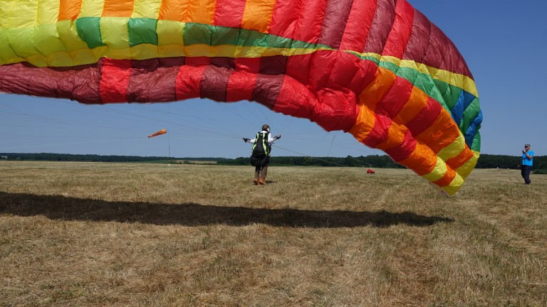 Ein Gleitschirmspringer steht auf einer grün-braunen Wiese und landet mit seinem Gleitschirm. Der Gleitschirm ist rot, orange, gelb, grün und blau. Rechts neben dem Gleitschirmspringer steht ein Mann. Es scheint ein blauer Himmel und im Hintergrund sind grüne Bäume zu erkennen.