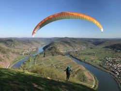 Ein Gleitschirmspringer ist in der Luft, sein Gleitschirm ist gelb und orange. Unter ihm sind Berge, grüne Wiesen, grüne Bäume, Häuser und Wasser zu sehen.