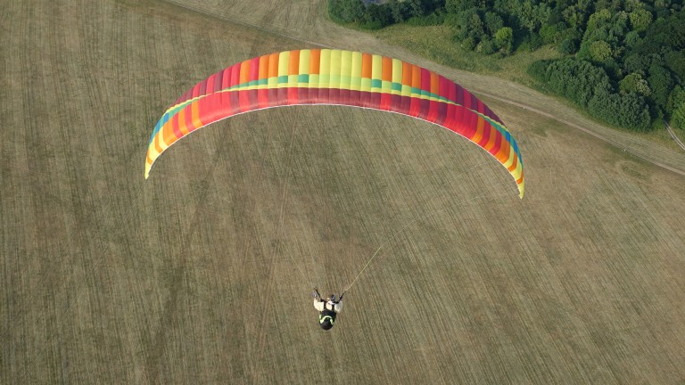 Ein Gleitschirmspringer ist in der Luft, sein Gleitschirm ist rot, orange, gelb, grün und blau. Unter ihm ist eine grün-braune Wiese. Oben rechts in der Ecke sind grüne Bäume zu erkennen.