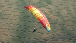 Ein Gleitschirmspringer ist in der Luft, sein Gleitschirm ist rot, orange, gelb, grün und blau. Unter ihm ist eine grün-braune Wiese. Oben rechts in der Ecke sind grüne Bäume zu erkennen.