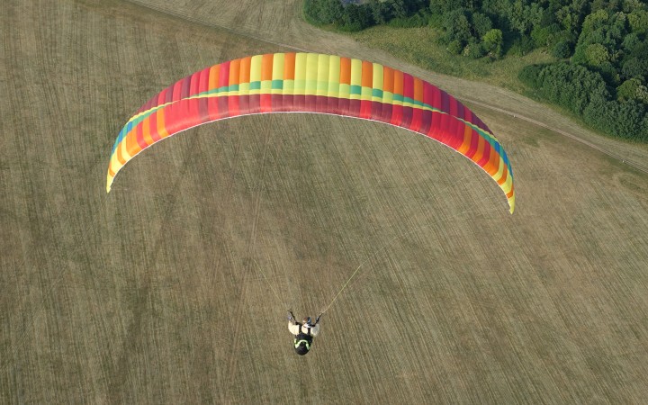Ein Gleitschirmspringer ist in der Luft, sein Gleitschirm ist rot, orange, gelb, grün und blau. Unter ihm ist eine grün-braune Wiese. Oben rechts in der Ecke sind grüne Bäume zu erkennen.