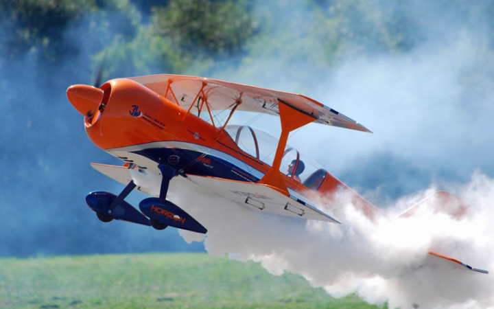 Orangener Modellflieger hebt ab. Hinter dem Flieger ist eine Rauchwolke und der Flieger startet auf einer Wiese. Hinter dem Flieger ist ein Wald mit grünen Bäumen.