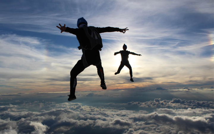 Zwei Menschen sind in der Luft mit Rucksäcken, wo Gleitschirme drin sind. Unter ihnen sind weiße Wolken und über ihnen ist ein blauer Himmel mit einem Schleier weißer Wolken.