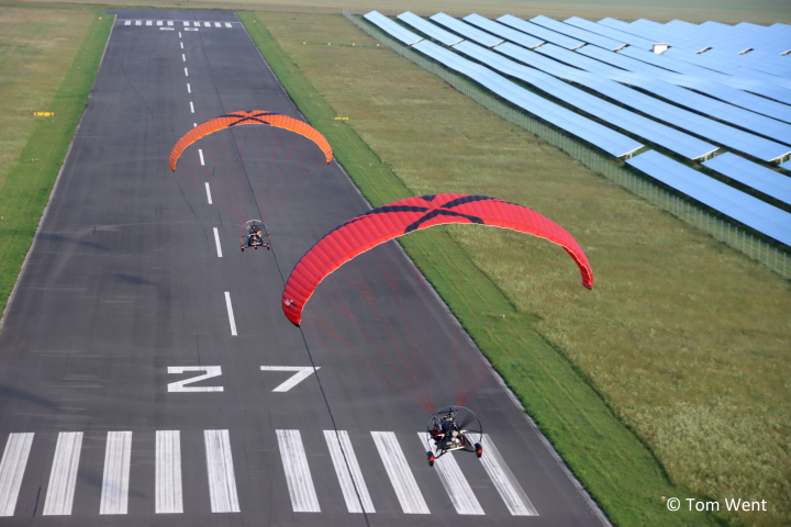 Zwei Ultraleichtflugzeuge die landen. Das Flugzeug hat einen orangenen Gleitschirm und das vordere Flugzeug hat ein roten Gleitschirm. Die Landebahn ist grau und hat weiße Markierungen. Neben der Landebahn ist eine grüne Fläche.