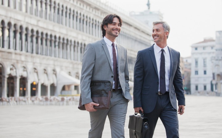 Zwei lächelnde Männer im Anzug und mit Aktentasche die über einen Platz laufen