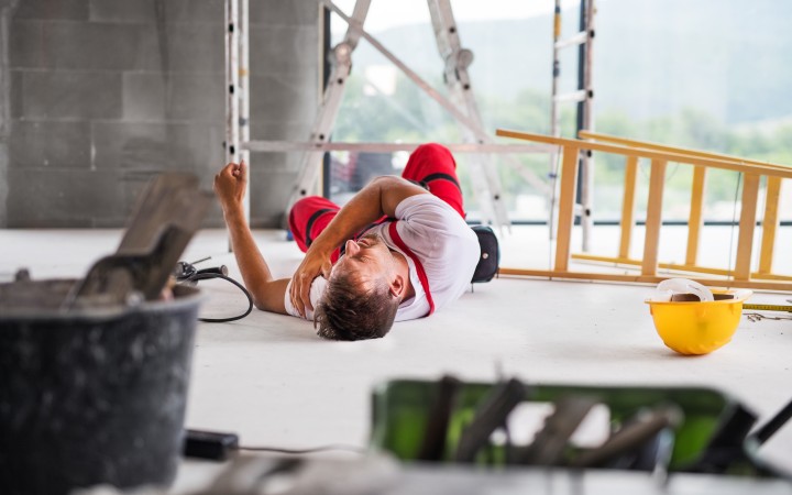 Mann in Arbeitshose leigt auf Boden. Man sieht eine umgekippte Leiter, neben ihm liegt ein gelber Schutzhelm, im Hintergrund ist eine weitere Leiter zu erkennen und ein Gerüst, im Vordergrund stehen andere Arbeitsutensilien