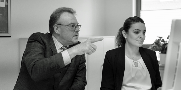 Thomas Sawade links und Mitarbeiterin rechts sitzen an einem PC, hinter Ihnen steht ein Regal und ein fenster. das bild ist schwarz-weiß.