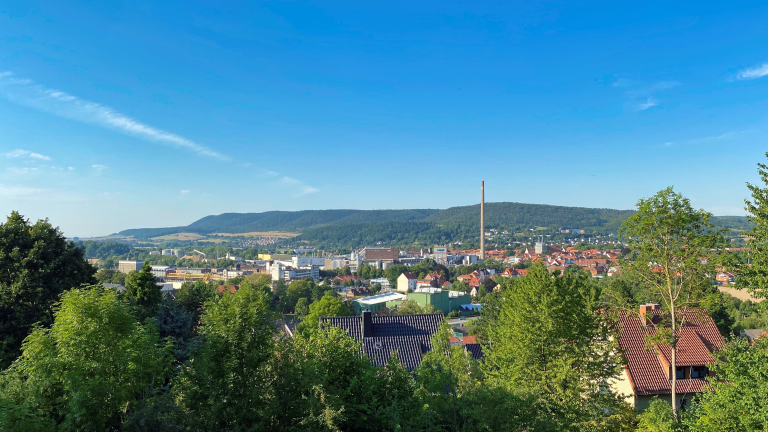 Man sieht viele Häuser, einen grauen Turm, viele grüne Bäume un deinen blauen Himmel