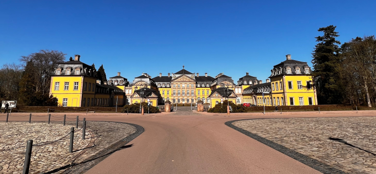 Gelbes Haus mit schwarzem Dach.Vor dem Haus ist ein Weg und neben dem Haus stehen Baüme und er Himmel ist blau