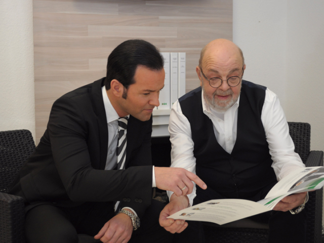 Timo Föhles links und ein Mitarbeiter rechts sitzen auf schwarzen Stühlen und gucken in eine Zeitschrift. Sie sitzen vor einer beigen Wand und man erkennt hinter Ihnen drei HDI Ordner auf einem weißen regal