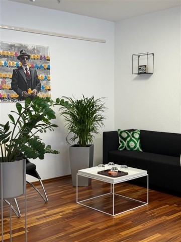 Rechts steht ein schwarzes Sofa, vor dem Sofa steht ein weißer Tisch. Links neben dem Sofa steht eine Pflanze. Links im Bild steht eine weitere Pflanze und hinter dieser Pflanze hängt ein Bild an der Wand. Auf dem Bild ist ein Mann zu erkennen.