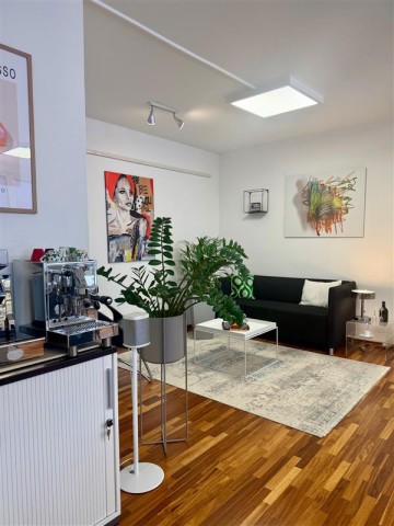 Eine schwarzes Sofa steht an einer weißen Wand. Vor dem Sofa liegt ein grauer Teppich auf dem ein kleiner weißer Tisch steht. Rechts neben dem Sofa steht ein Glastisch, auf dem eine Lampe steht und links neben dem Sofa steht eine grüne Pflanze mit einem weißen Topf. Über und neben dem Sofa hängen bunte Bilder. Links im Bild steht ein weißer Schrank, auf dem Schrank steht eine Kaffeemaschine und neben der Maschine steht eine Musikbox in weiß und eine Pflanze.