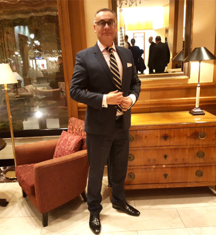 Mitarbeiter Wolf rainer Gust ist in einem Anzug. Im Hintergrund ist links von Ihm ein roter Sessel und ein Lampe und rechts von Ihm steht ein braunes Regal. Auf dem Regal steht eine Lampe und über dem Regal hängt ein Spiegel