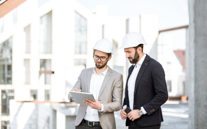 Zwei Architekten auf einem Bauobjekt, die sich unterhalten. Beide Herren tragen einen weißen Bauhelm und einen Anzug.