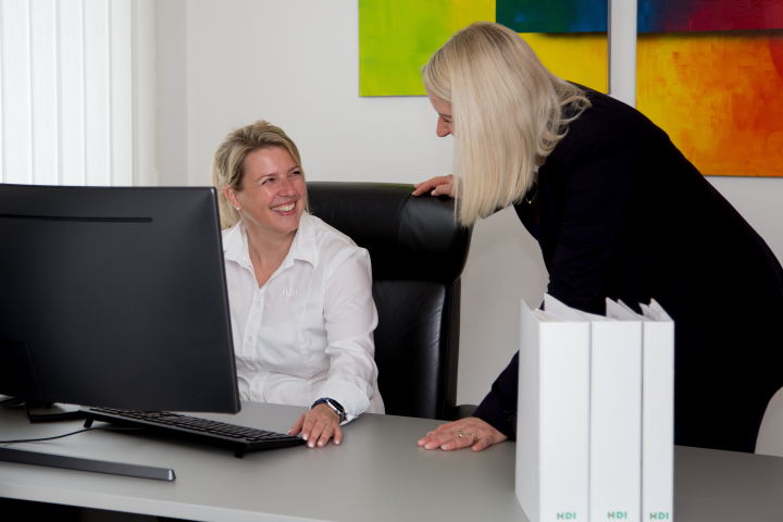 Yvonne Janowski sitzt auf einem schwarzen Stuhl am Schreibtisch auf dem ein PC und drei Ordner stehen. Rechts neben Ihr steht eine Mitarbeiterin, die nur von der Seite zu sehen ist. Hiter Ihnen  hängt ein buntes Bild.